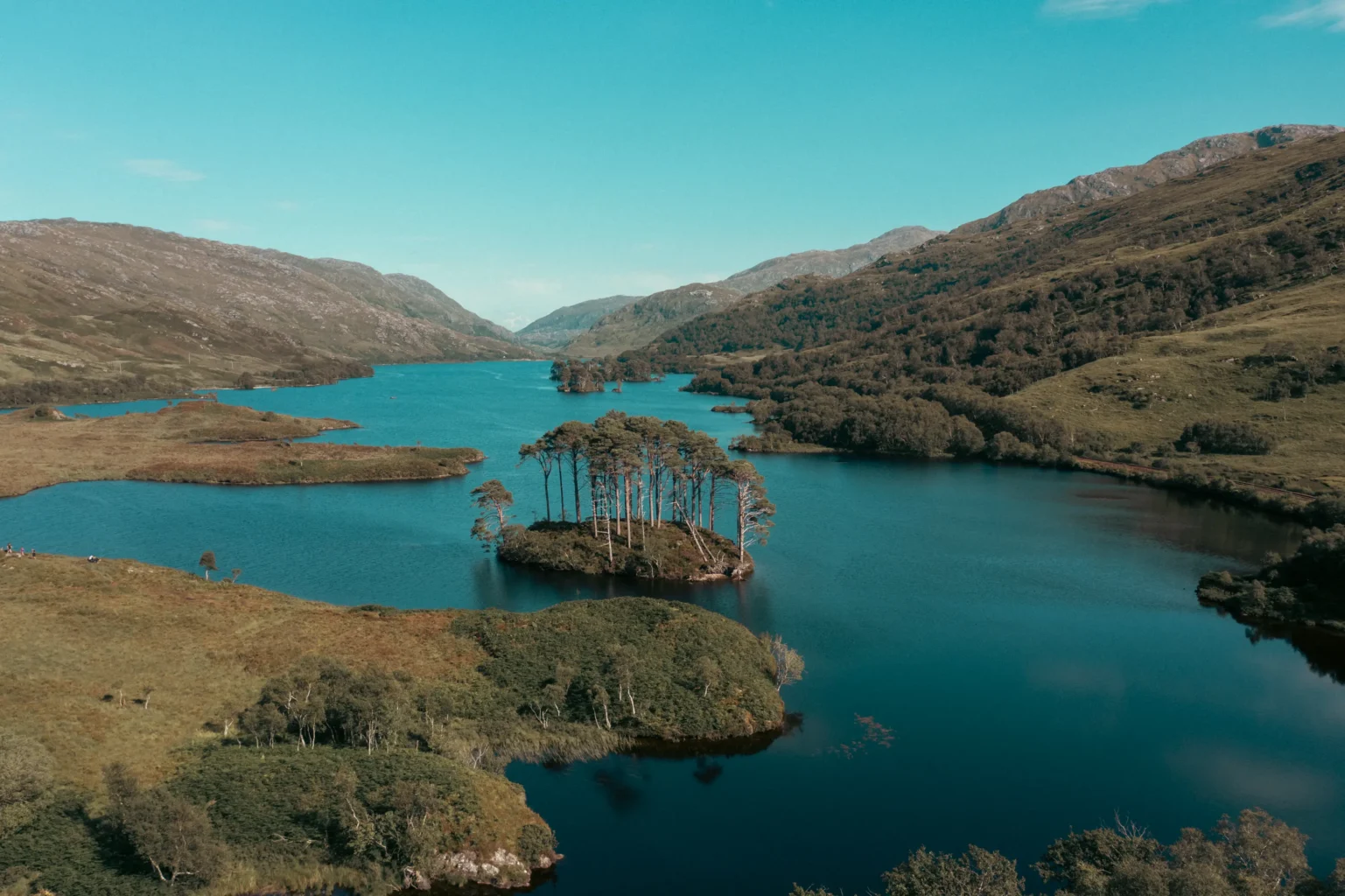 Aerial drone shot of Loch Eilt in the Scottish Highlands — small pine-covered island surrounded by vivid blue water and rolling hills under a clear sky, captured by OV Production
