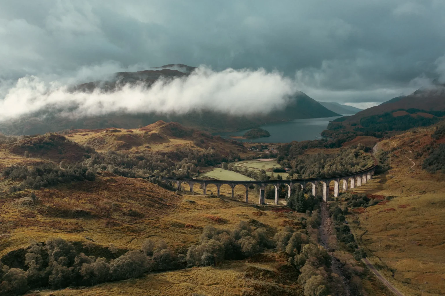 Wide aerial drone shot of Glenfinnan Viaduct curving through the Scottish Highlands in autumn, with Loch Shiel and low cloud-covered mountains in the background — aerial cinematography by OV Production