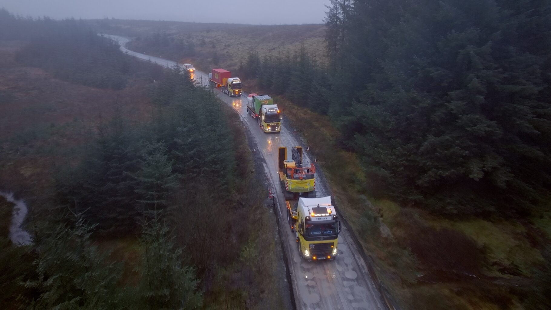 Aerial drone shot of a heavy haulage convoy carrying abnormal loads along a remote Highland road through Scottish moorland and forestry in wet weather — industrial logistics filming by OV Production