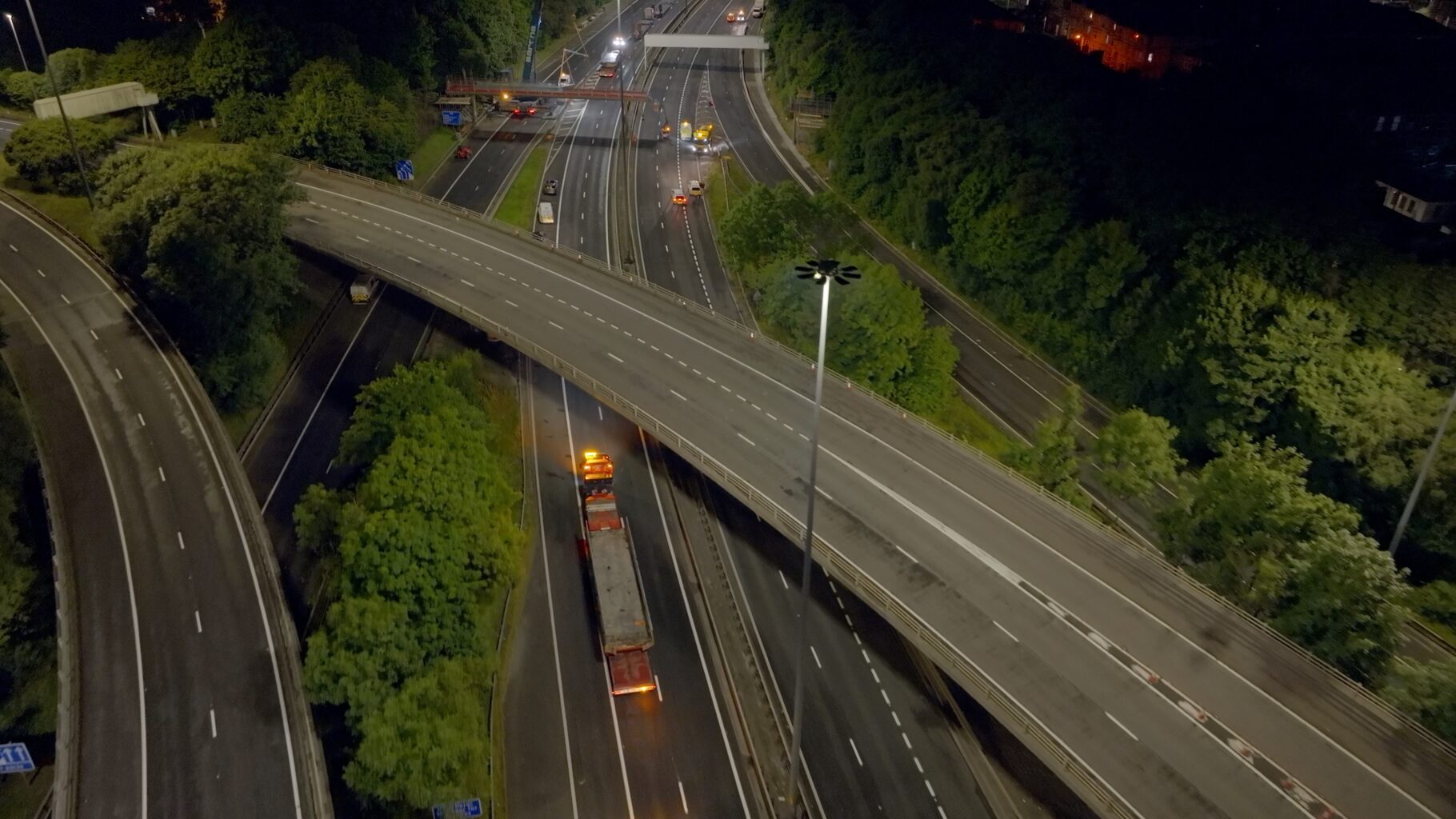 Night aerial drone shot of an Amey road maintenance operation — heavy haulage lorry passing under a motorway bridge during overnight infrastructure works, filmed by OV Production Scotland