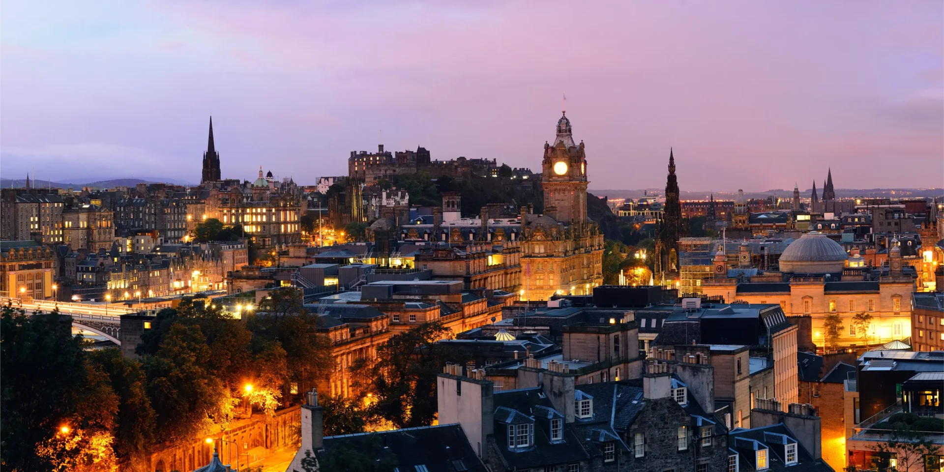Edinburgh city skyline at dusk photographed from Calton Hill — Balmoral Hotel clock tower, Edinburgh Castle, and the Scott Monument lit up against a purple evening sky, captured by OV Production